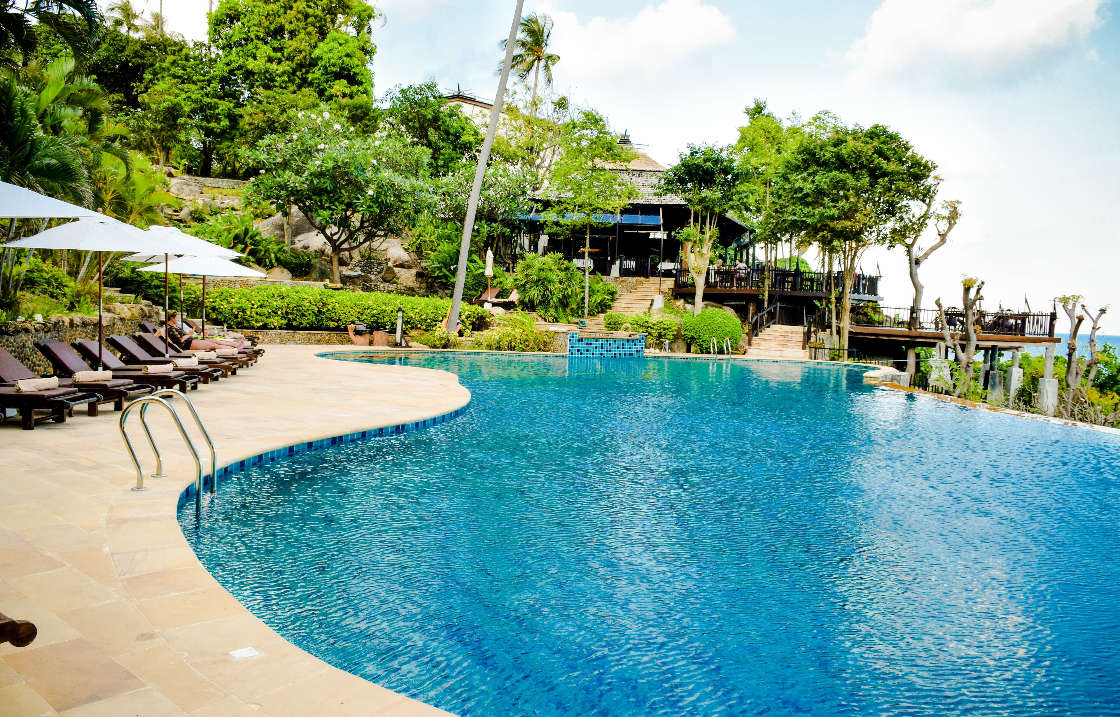 A pool area with lounge chairs and umbrellas, surrounded by lush greenery and a building with a balcony.