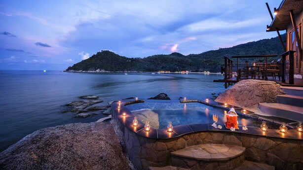 Piscina de piedra infinita con velas y una linterna, con vistas al mar tranquilo y un paisaje montañoso.