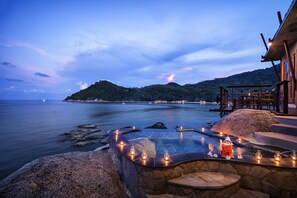 Piscina de piedra infinita con velas y una linterna, con vistas al mar tranquilo y un paisaje montañoso.