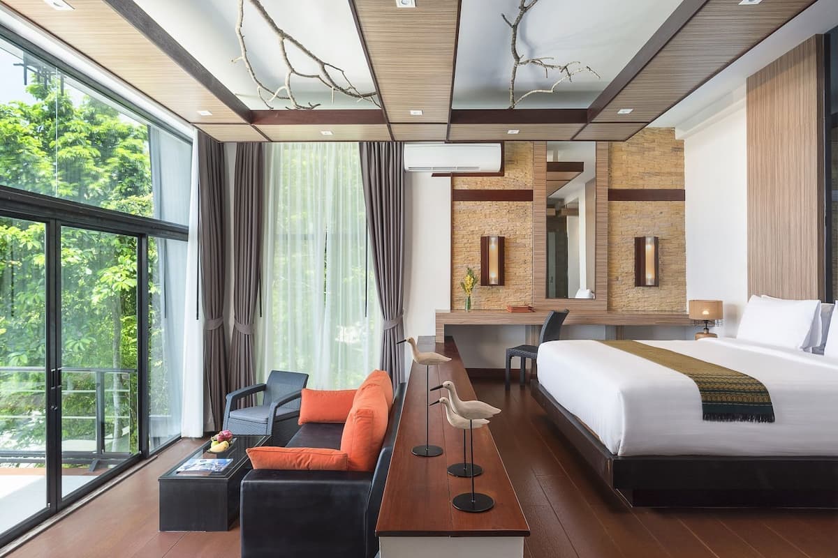 A modern hotel room with a large bed, a desk, a sofa, and a view of greenery outside.