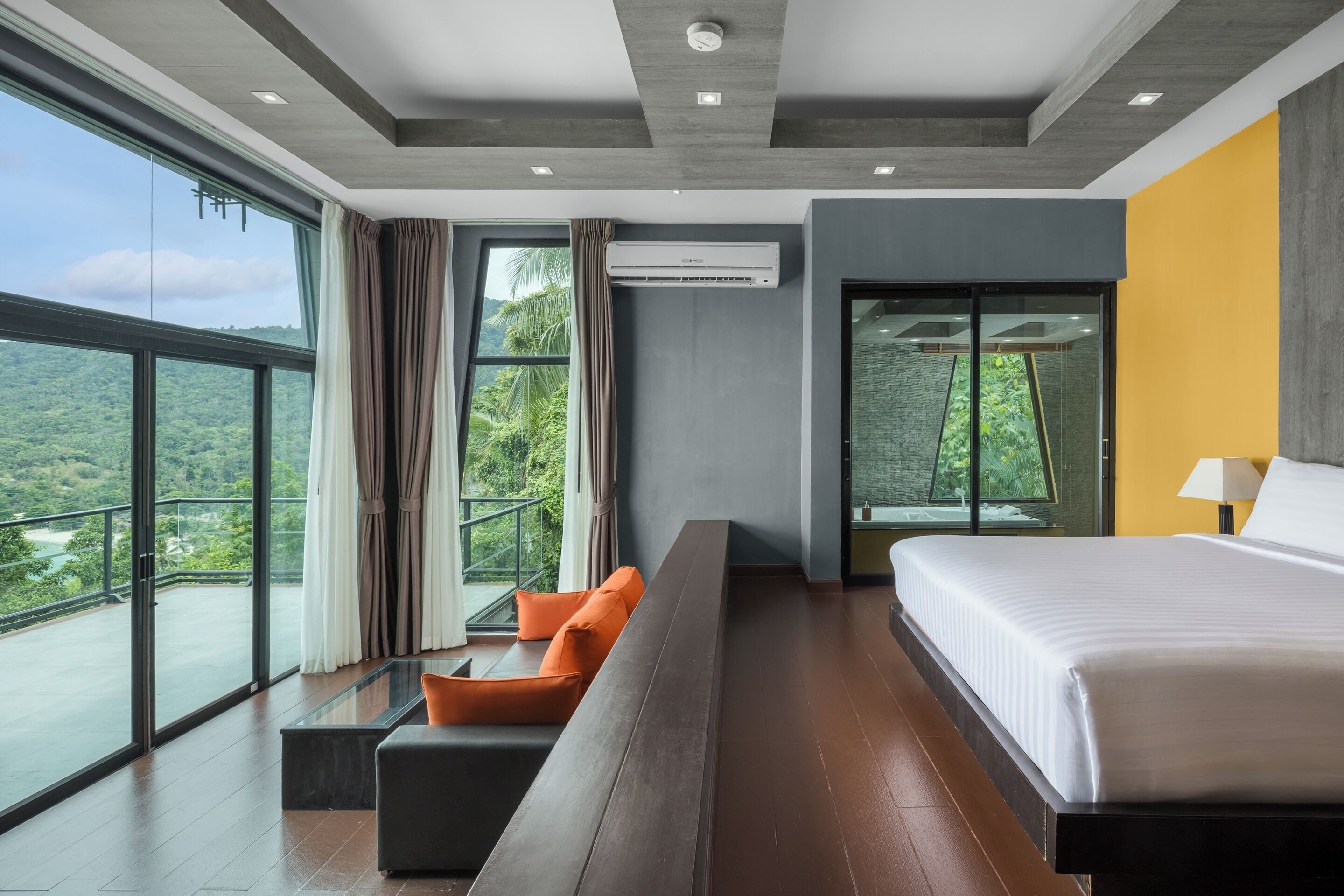 A modern hotel room with a large bed, a seating area with orange cushions, a glass table, and a view of greenery through large windows.