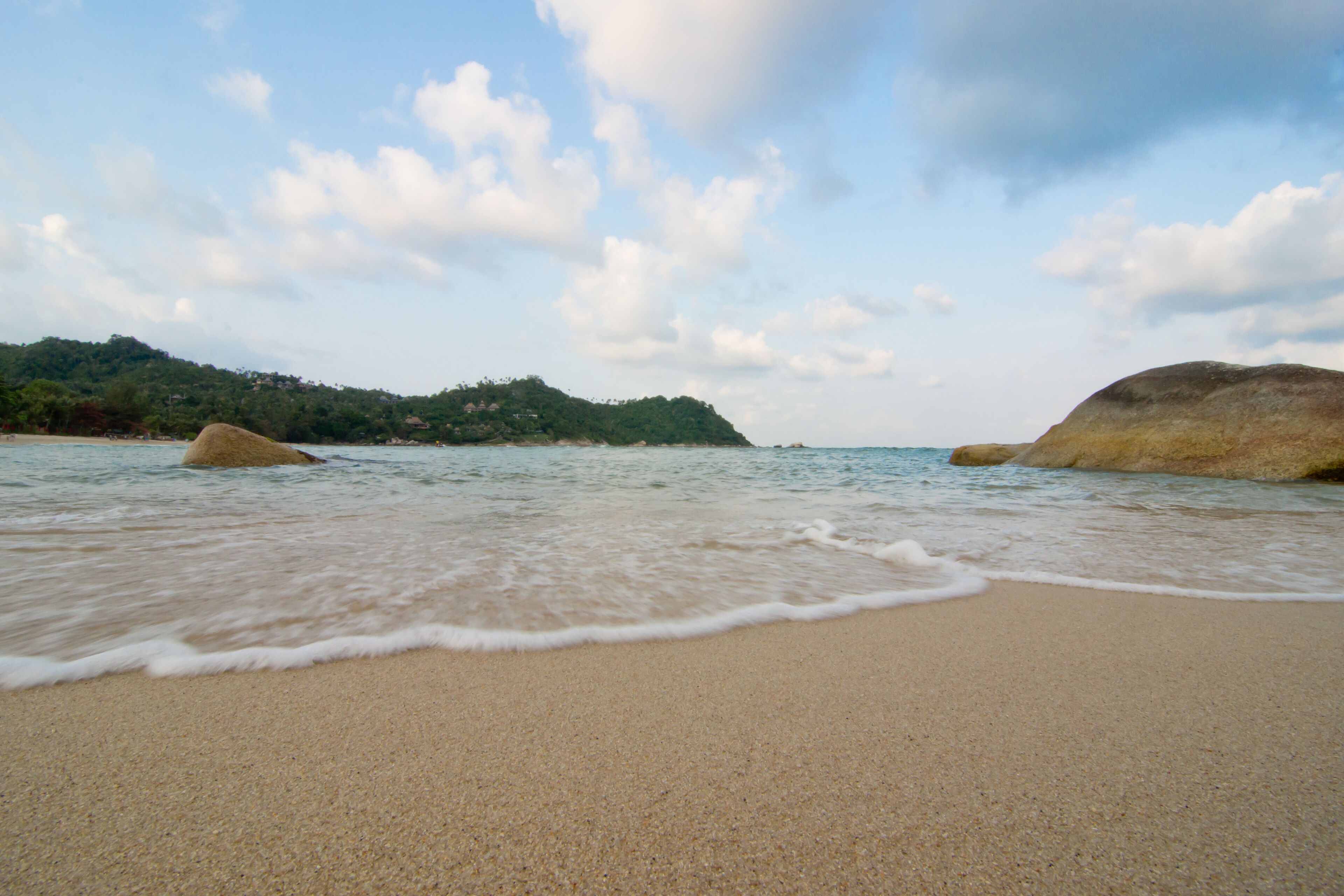 A beach with calm waves, a sandy shore, and a distant hill with trees.