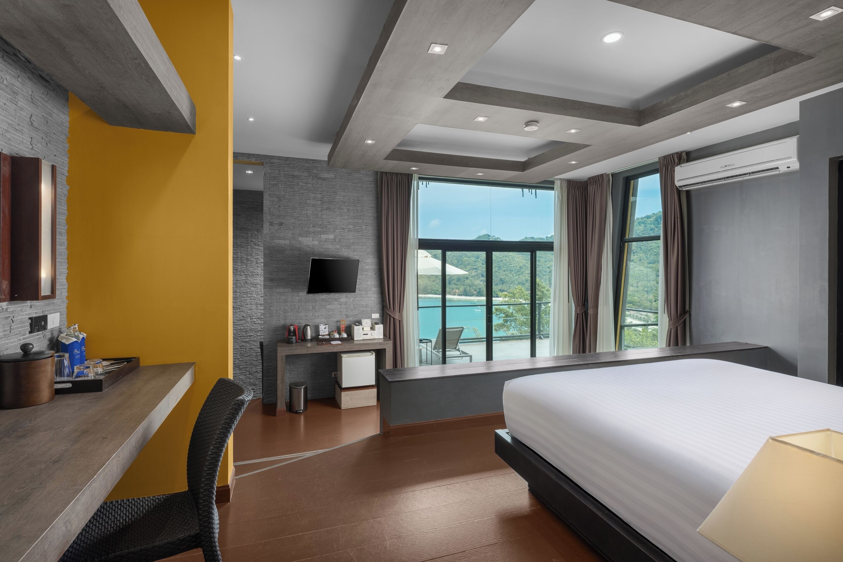 Een moderne hotelkamer met een groot bed, een bureau met stoel, een kleine kitchenette, een flatscreen-tv en een balkon met uitzicht op zee.