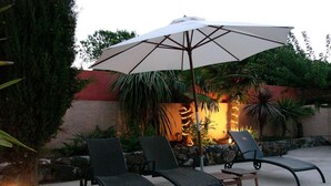 An outdoor seating area with three lounge chairs and a large umbrella, surrounded by tropical plants and a stone wall. - Hostellerie La Bergerie du Cap (Agde)