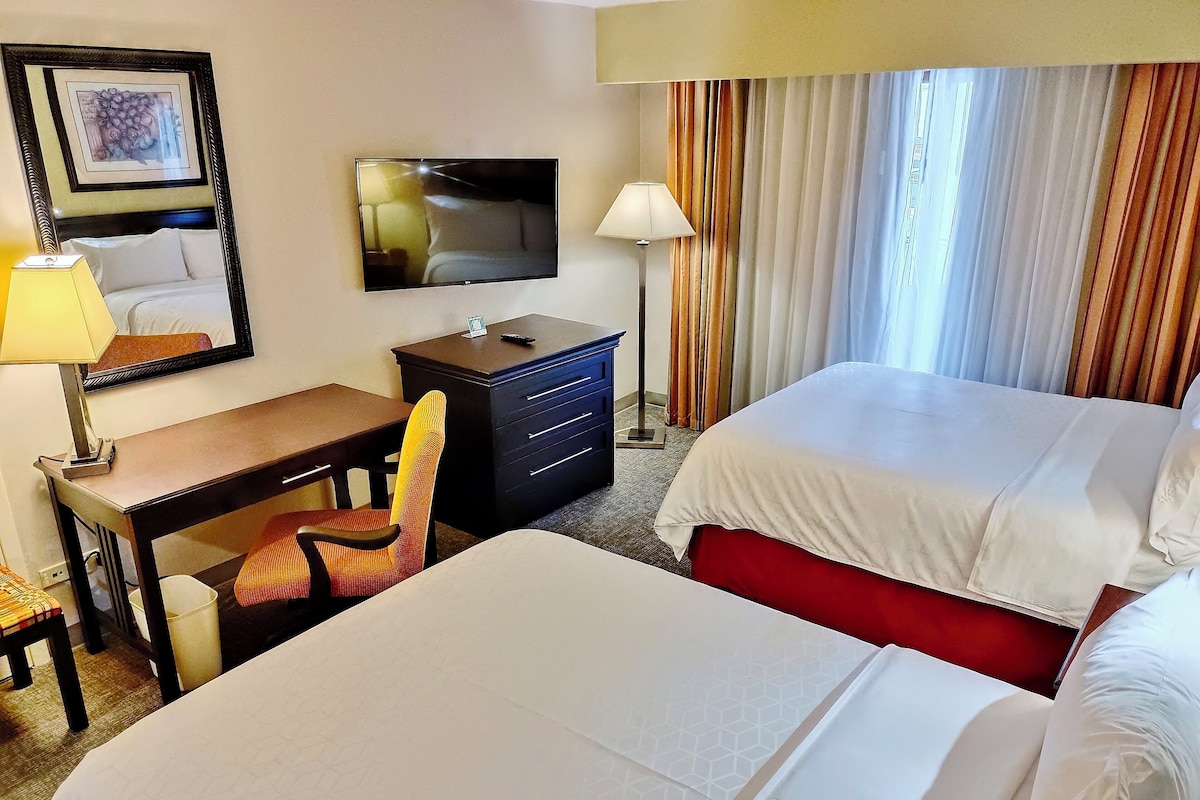 Quarto de hotel com duas camas, uma escrivaninha, uma cadeira, uma televisão e uma janela com cortinas.