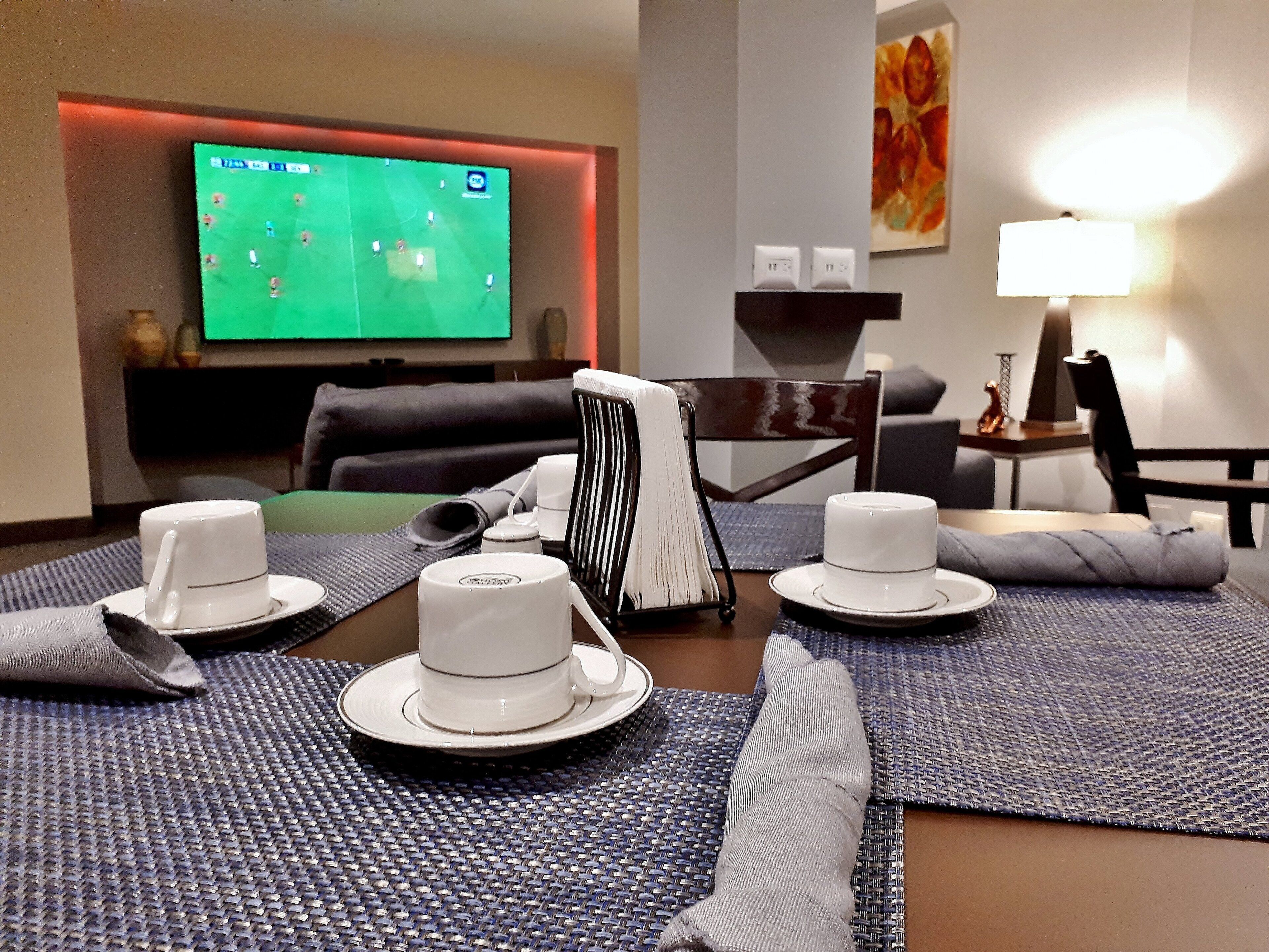 A hotel room with a dining table set for two, a flat-screen TV displaying a sports game, a sofa, and a painting on the wall.