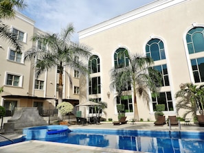 A pool area with lounge chairs and a small table, surrounded by palm trees and a building with large windows.