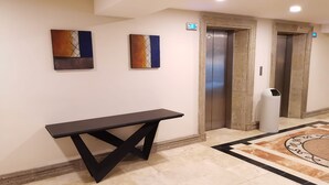 Un vestíbulo con piso de mármol, una mesa negra, dos cuadros abstractos, un ascensor y un cesto de basura.