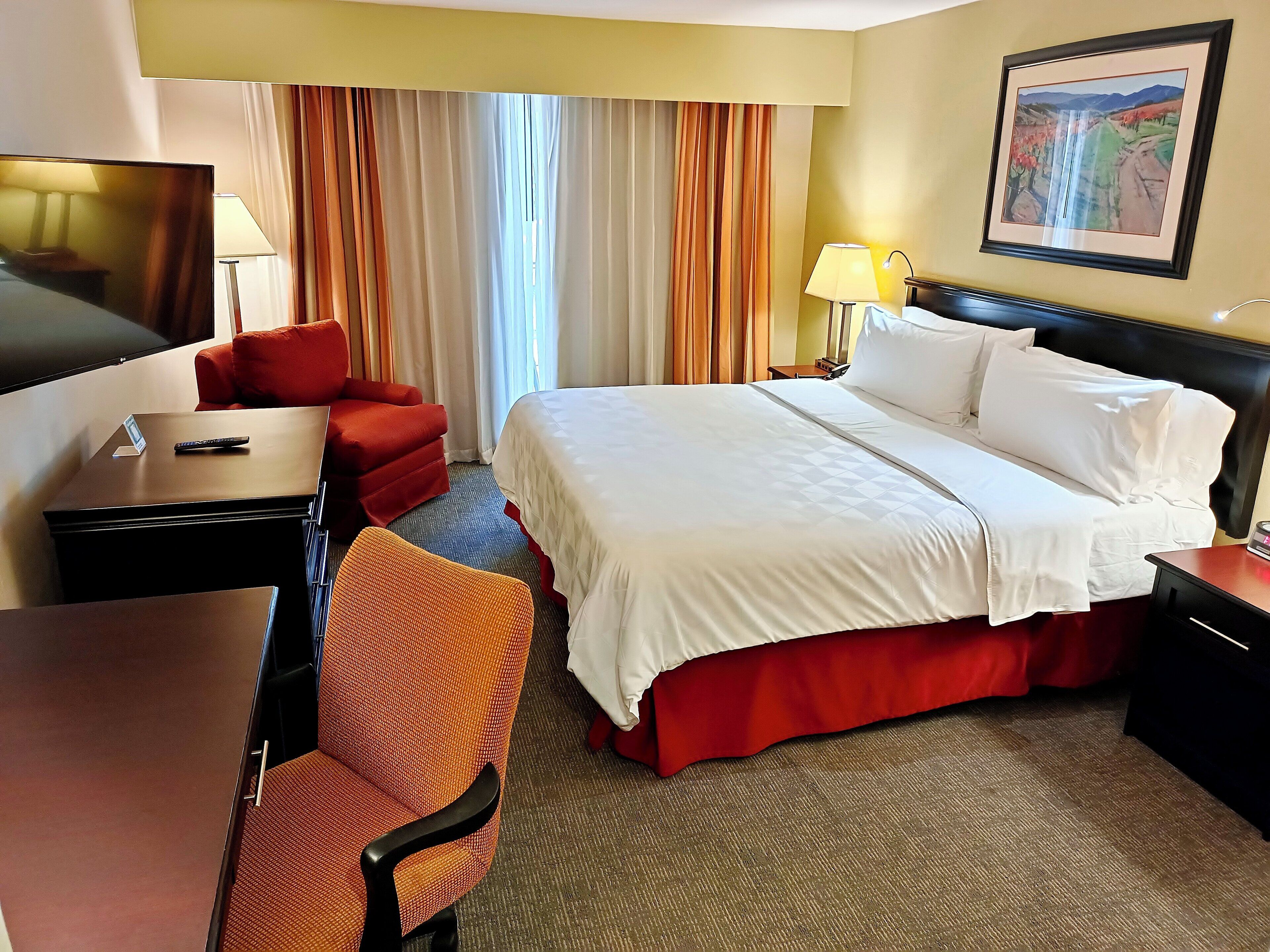 A hotel room with a bed, a desk, a chair, a TV, and a painting on the wall.