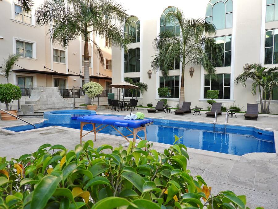 Una piscina con sillas de descanso y un sillón de masaje, rodeada de palmeras y un edificio con ventanas amplias.