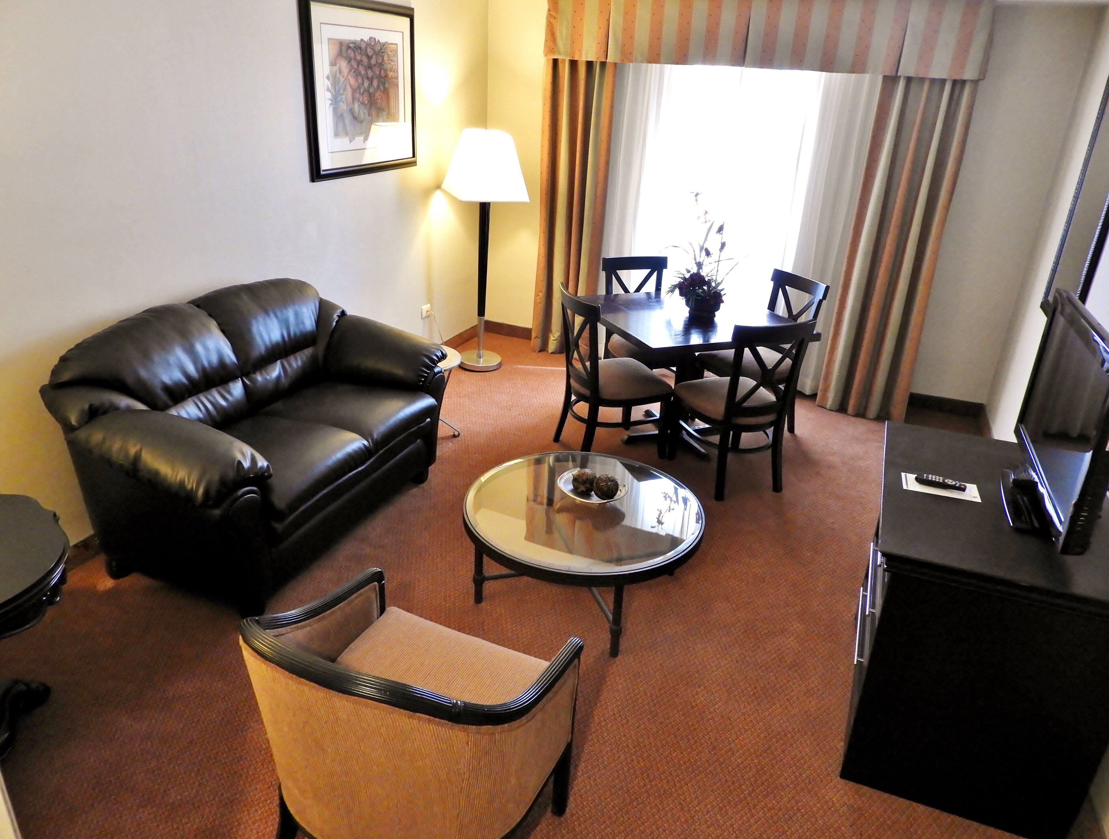 A hotel room with a leather sofa, a small round table, a wooden chair, a TV stand, and a lamp.