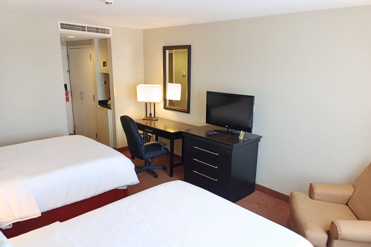 Quarto de hotel com duas camas, uma escrivaninha com computador, uma televisão e uma cadeira.