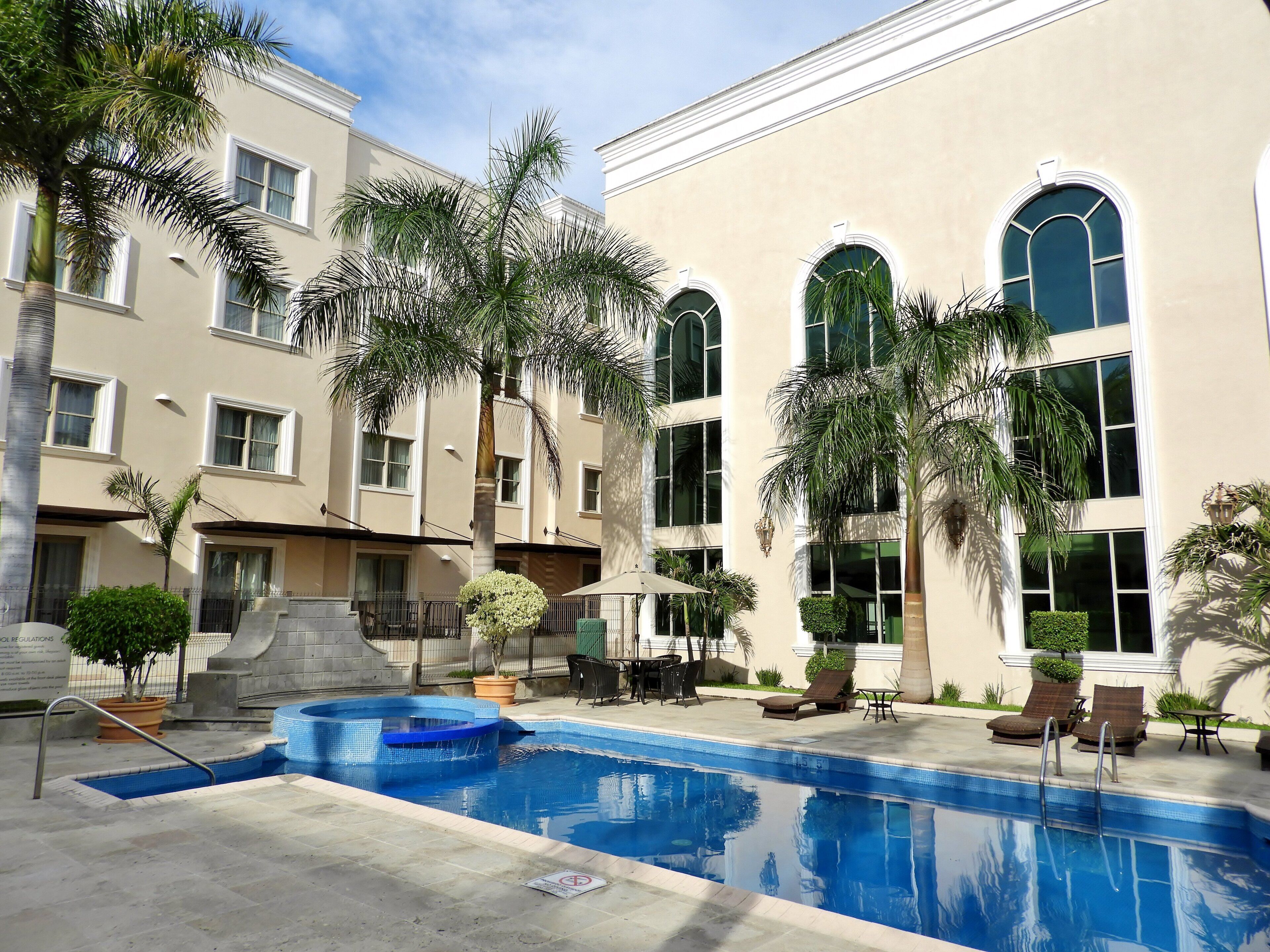 Une piscine avec un jacuzzi, entour ée de palmiers et de chaises longues, avec un bâtiment doté de grandes fenêtres en arrière-plan.