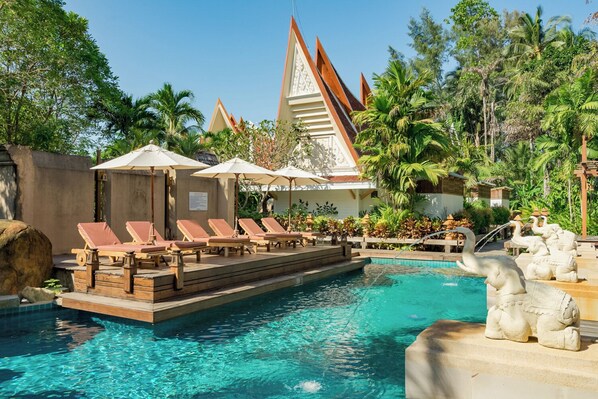 Un área de piscina con sillones de descanso y sombrillas, rodeada de vegetación tropical y arquitectura tradicional.