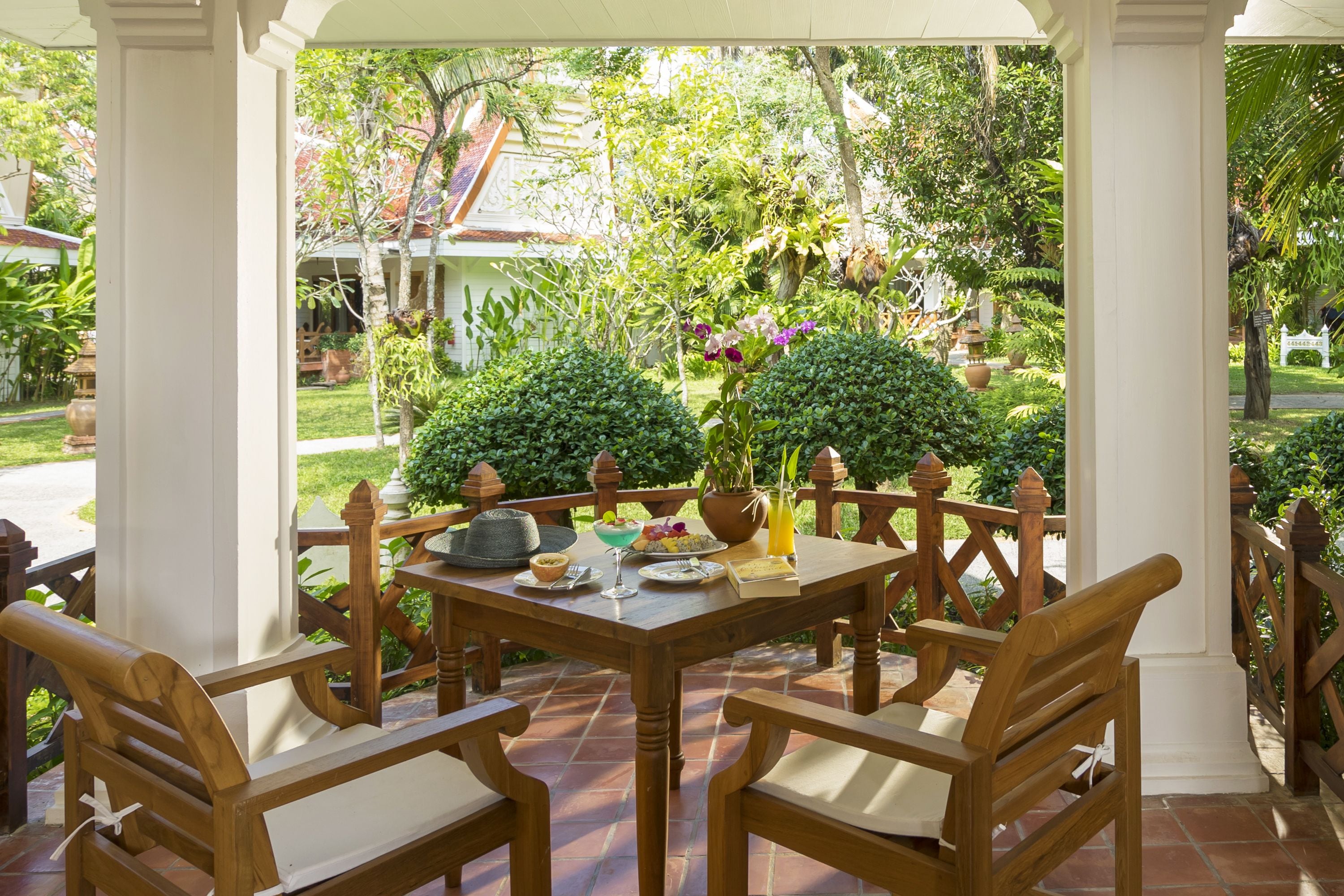 A wooden table set with plates, a bowl, and a glass of juice, surrounded by chairs, with a view of a garden and a building in the background.