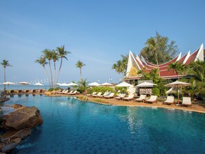 A pool area with lounge chairs and umbrellas, palm trees, and a building with a distinctive roof.