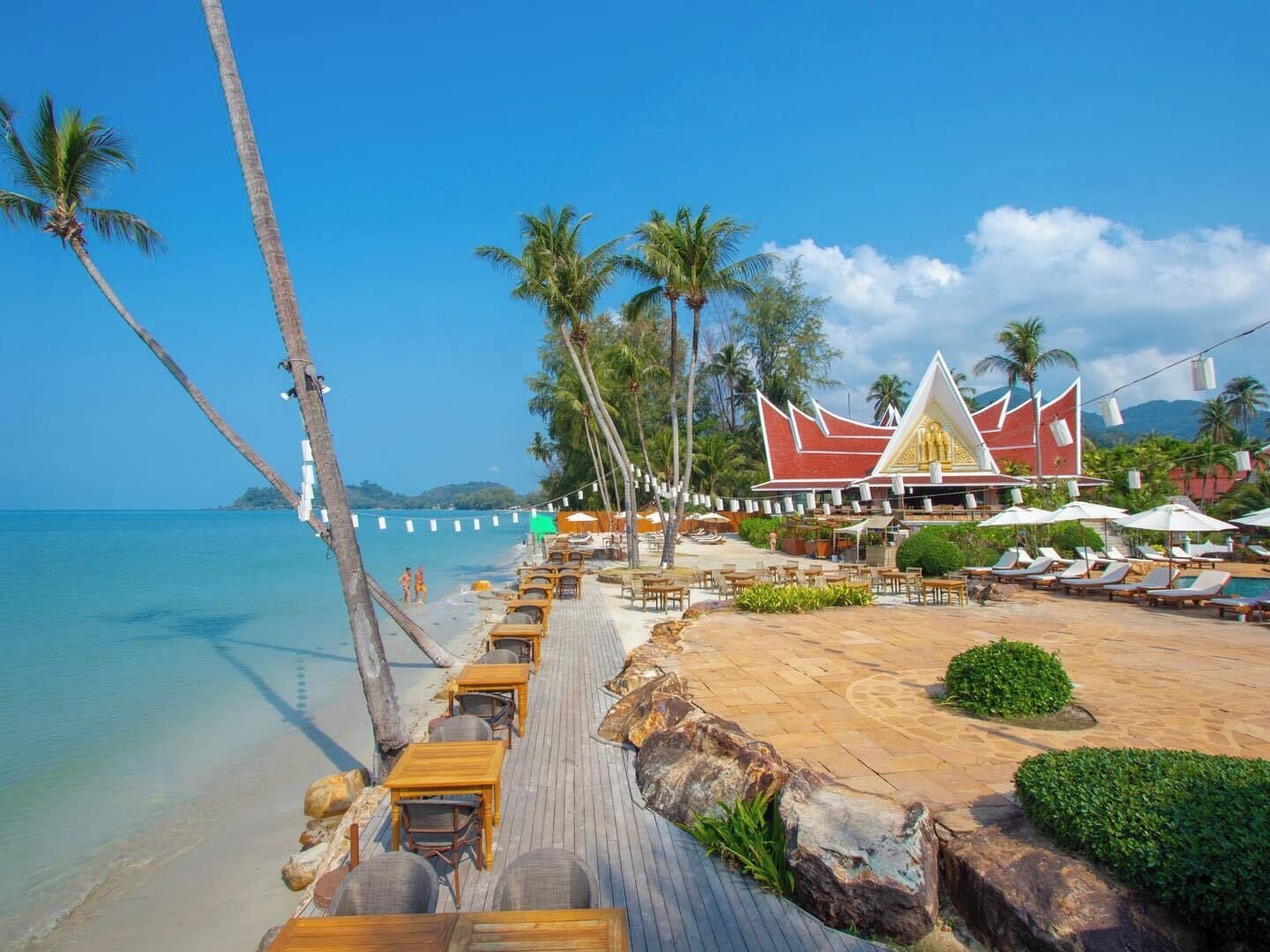 Un resort frente al mar con una terraza de madera, sillones de descanso y un edificio tradicional de techo puntiagudo.
