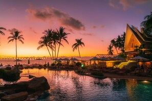 A resort pool area with palm trees, lounge chairs, and a sunset view.