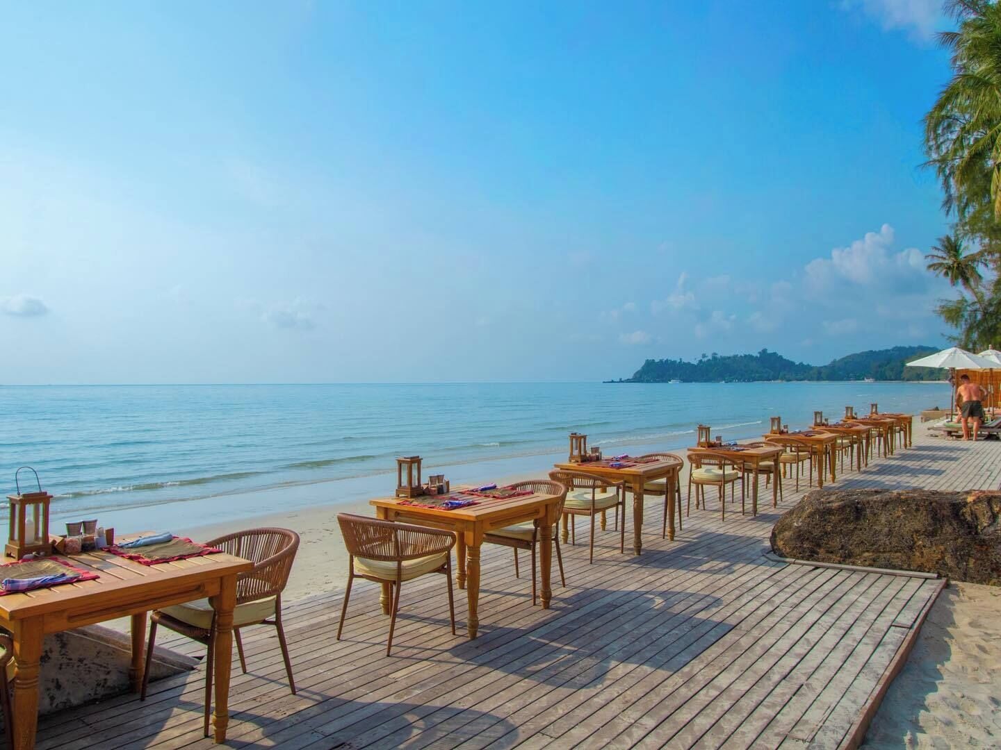 A wooden deck with tables and chairs overlooking a calm sea, with a distant island visible.