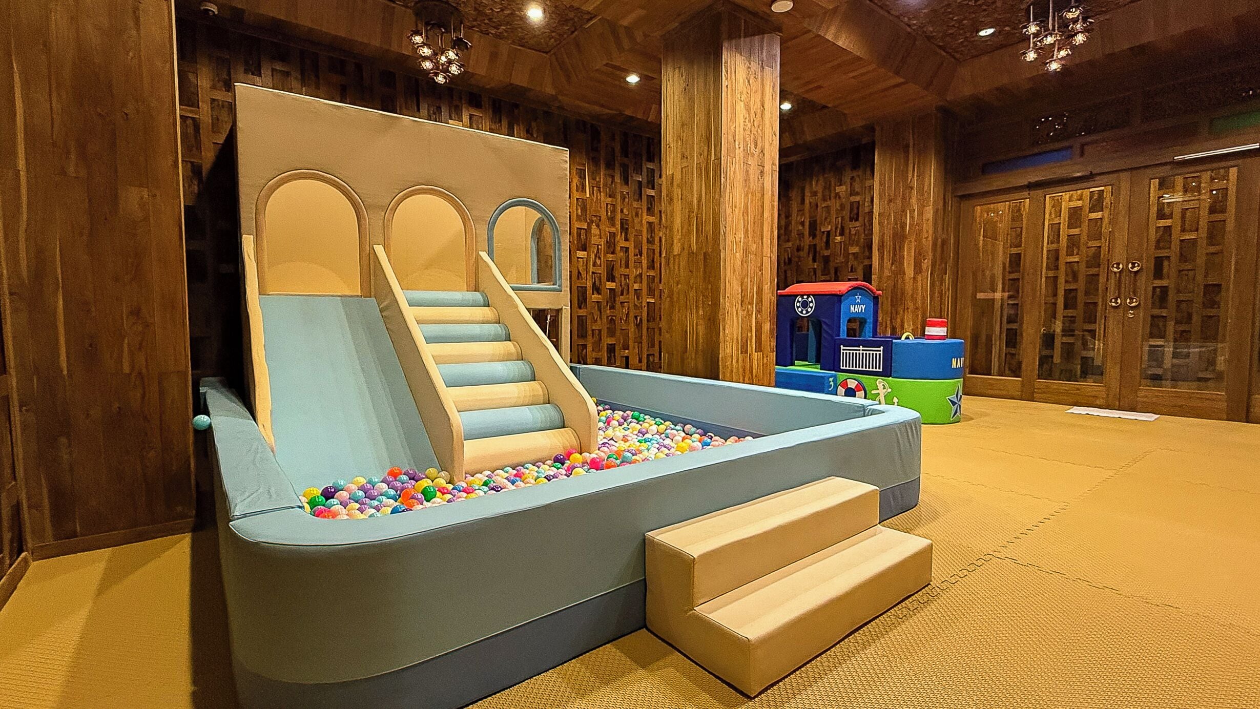 Uma área de recreação com escorregador, piscina de bolinhas, paredes de madeira e um trem de brinquedo.