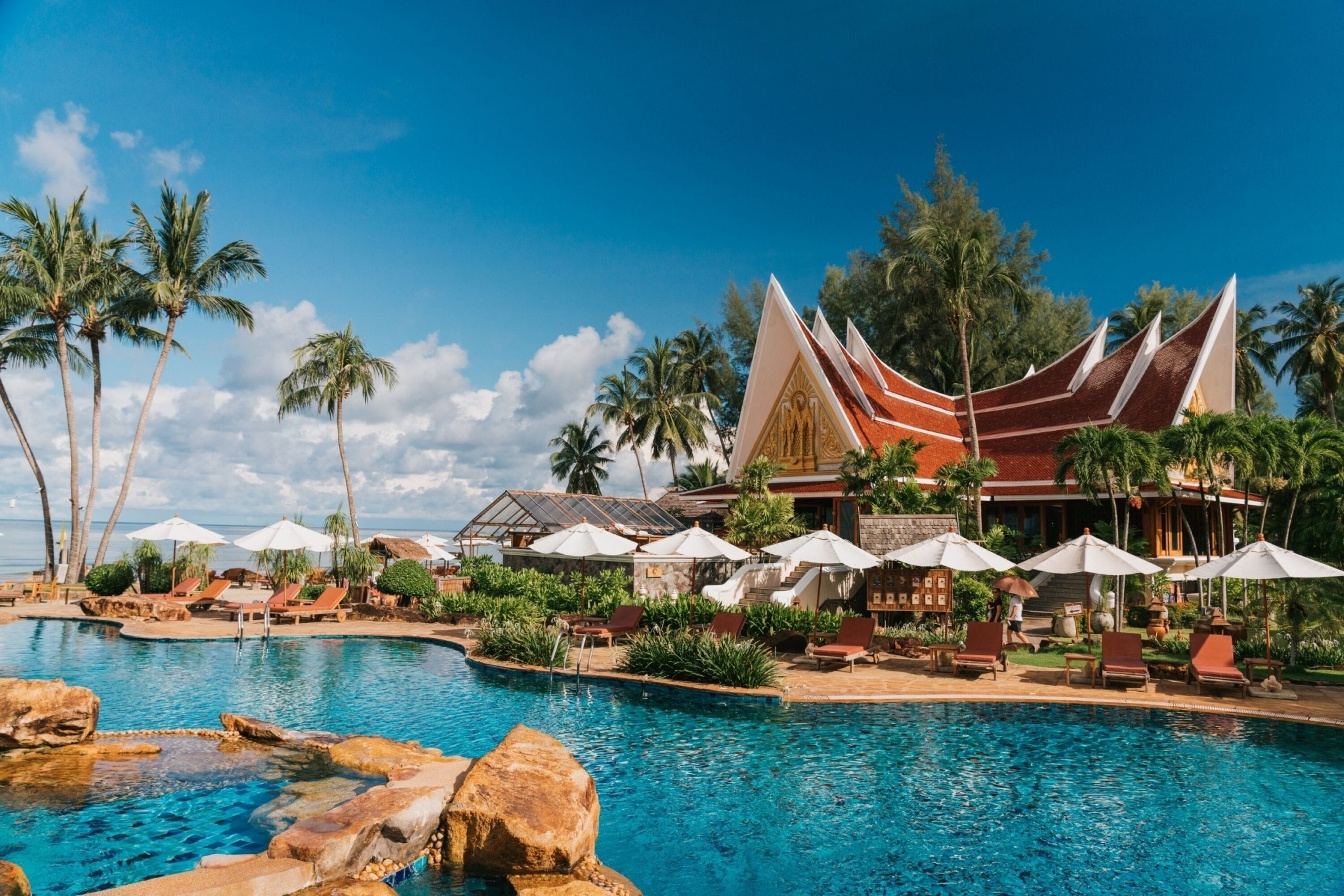 A resort with a swimming pool, lounge chairs, umbrellas, and palm trees.