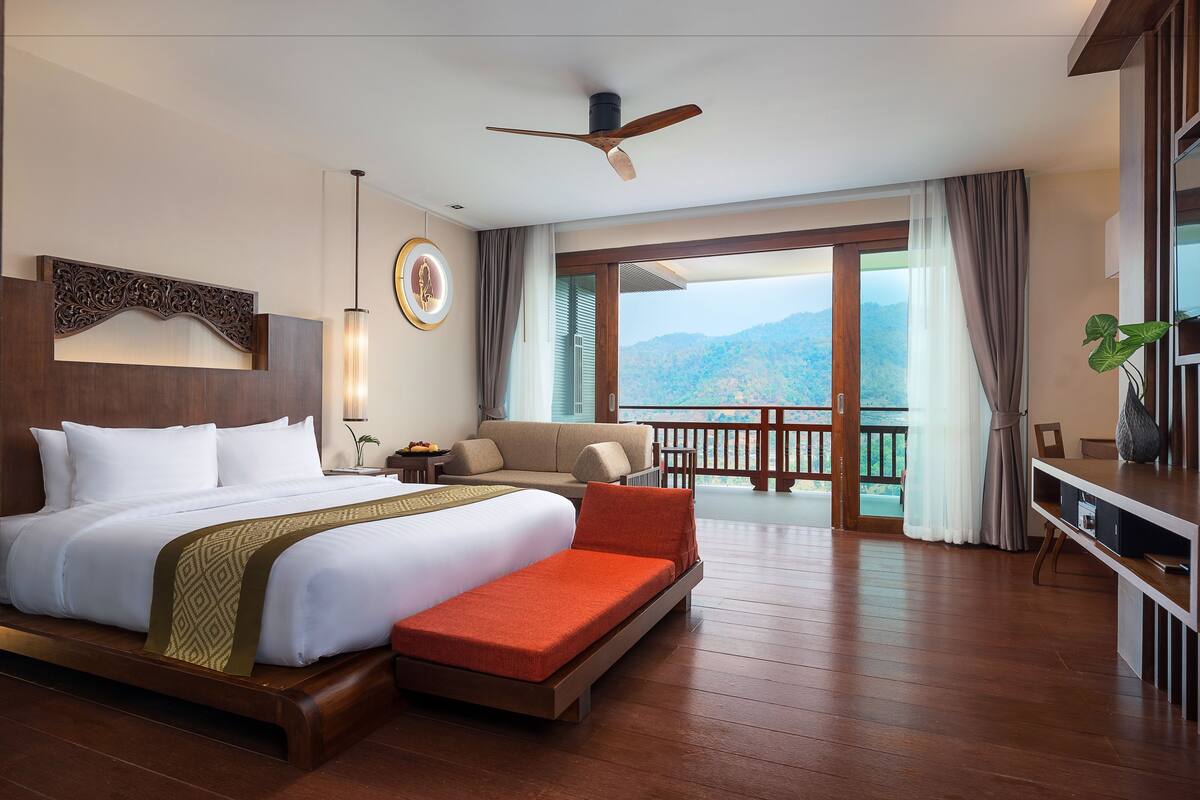 Quarto de hotel com uma cama grande, um sofá, uma varanda com vista para as montanhas e um ventilador de teto.