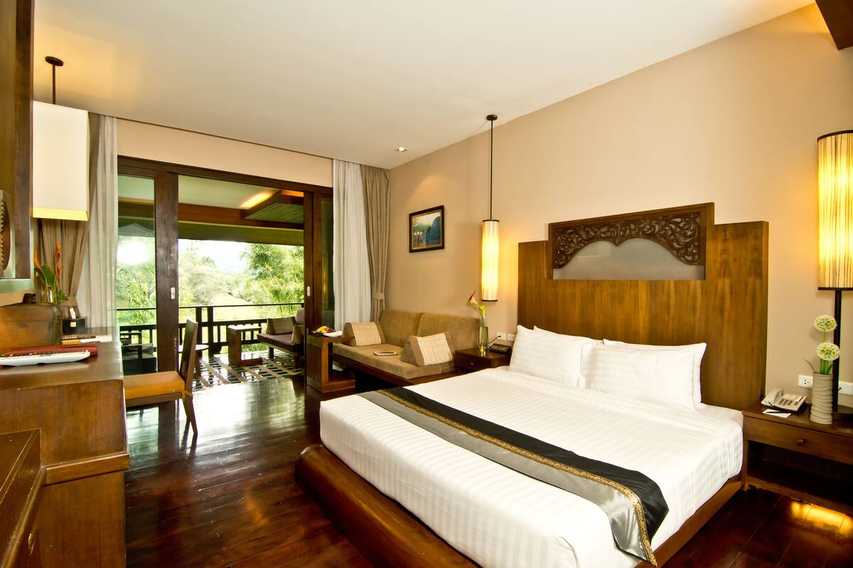 Quarto de hotel espaçoso com uma cama grande, cabeceira de madeira, uma mesa com telefone e vista para o exterior por uma porta de correr.