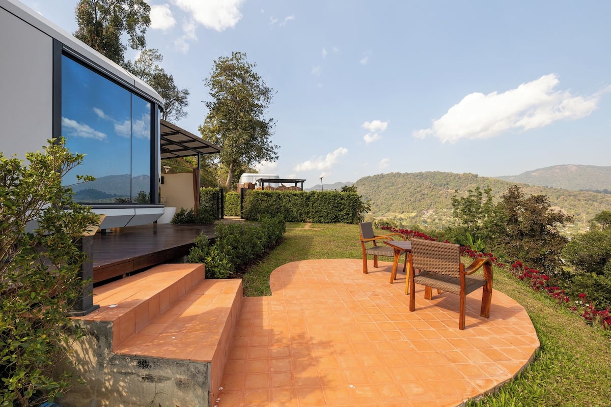 Um pátio com um banco e uma mesa, rodeado por vegetação e vista para colinas.