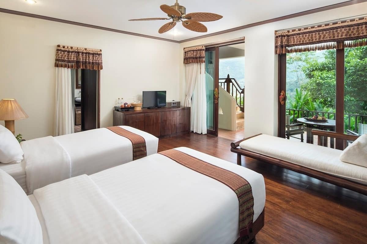 Quarto de hotel com duas camas, televisão, escrivaninha e varanda com vista para o verde.