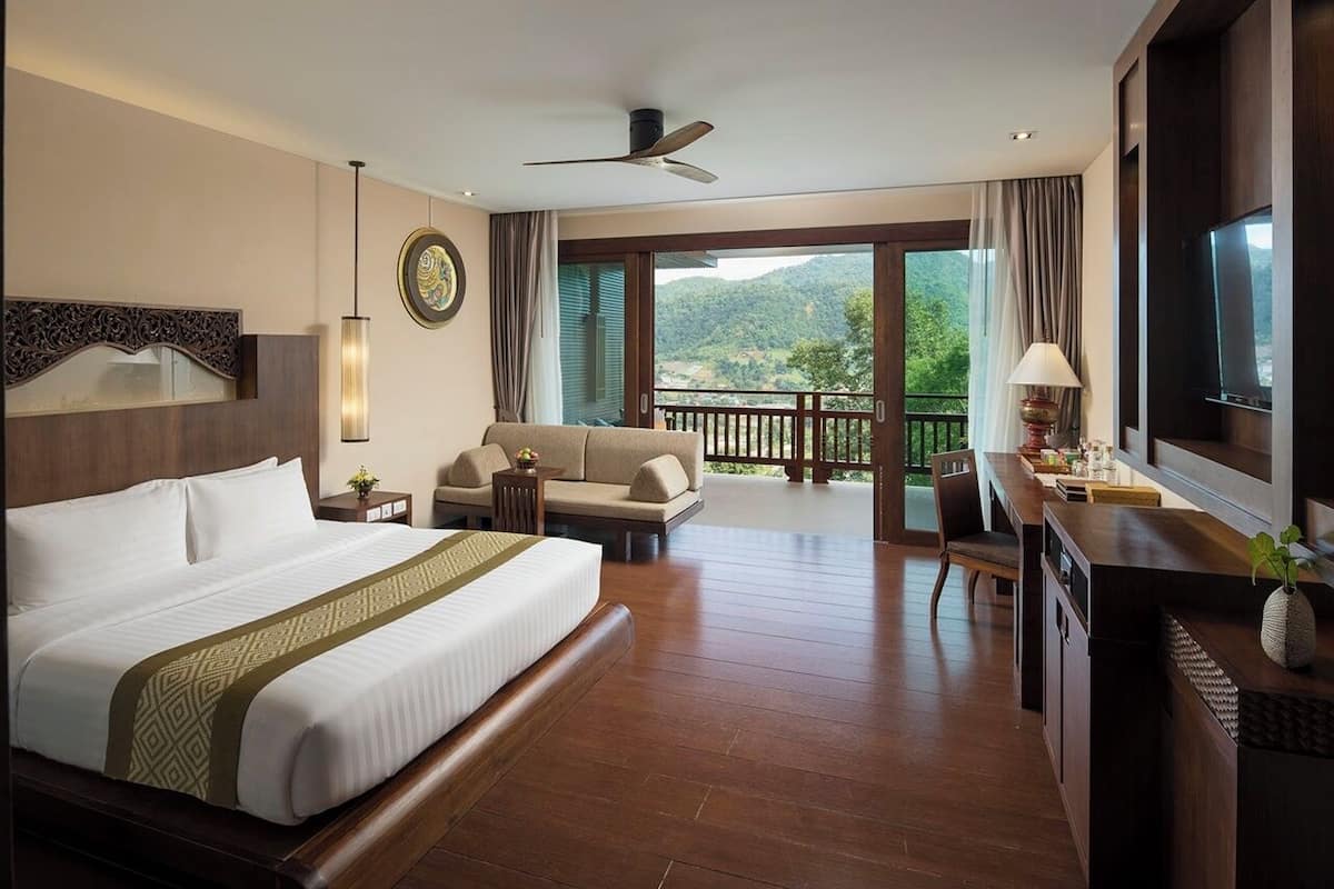 Quarto de hotel com uma cama grande, um sofá, uma mesa de trabalho e uma varanda com vista para as montanhas.