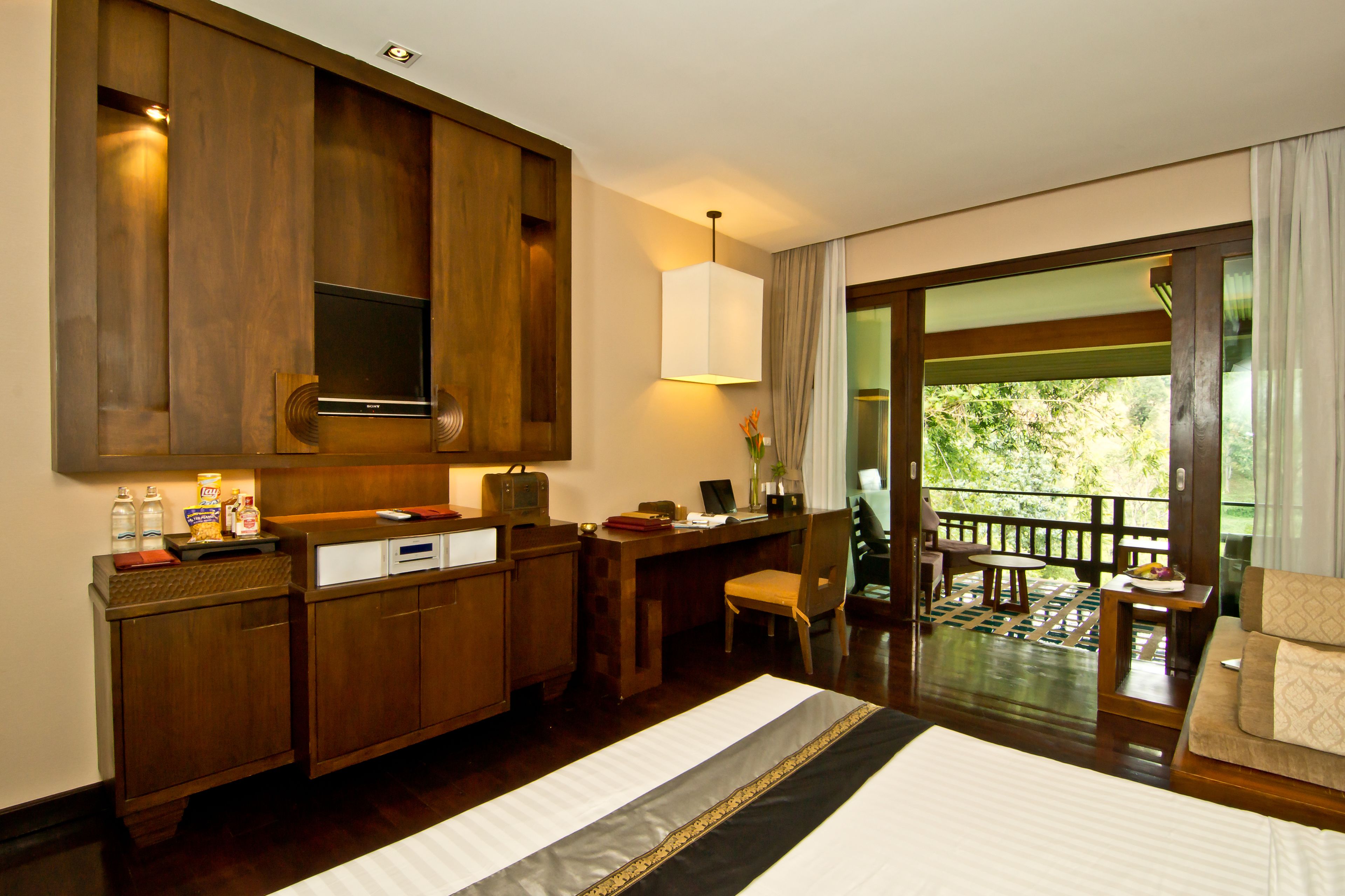 Quarto de hotel com uma cama grande, móveis de madeira, uma mesa com computador, televisão e varanda com vista para o verde.
