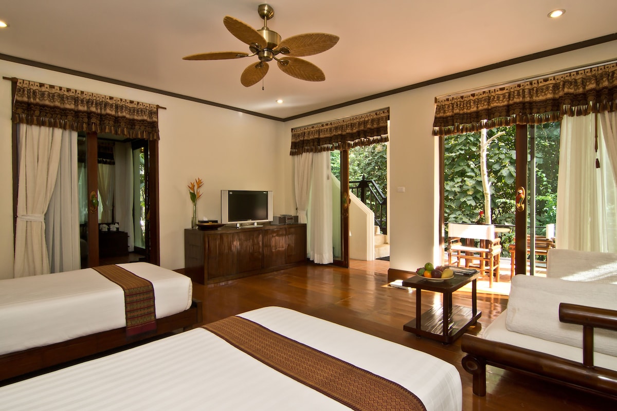 A hotel room with two beds, a TV, a desk with a fruit bowl, and a balcony with a view of greenery.