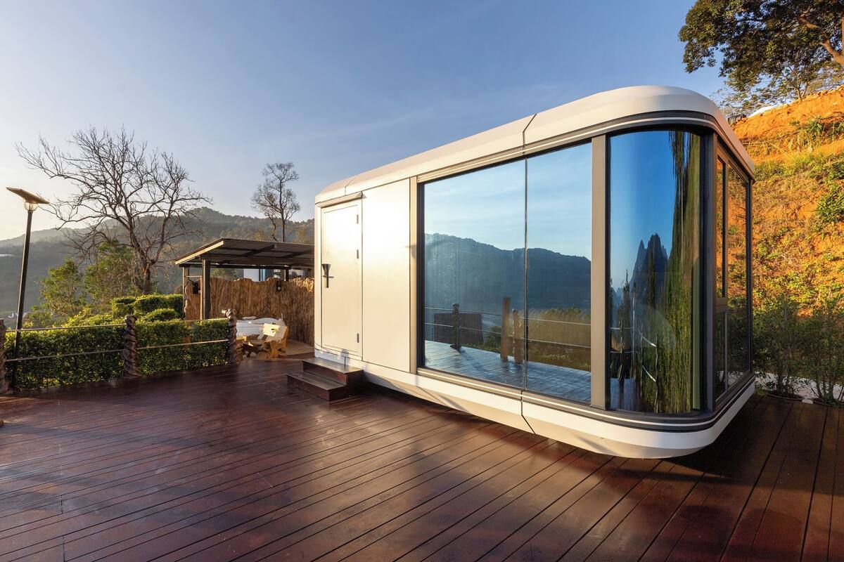 Estrutura moderna de vidro curvo com deck de madeira, rodeada por paisagens naturais e vista clara das montanhas.
