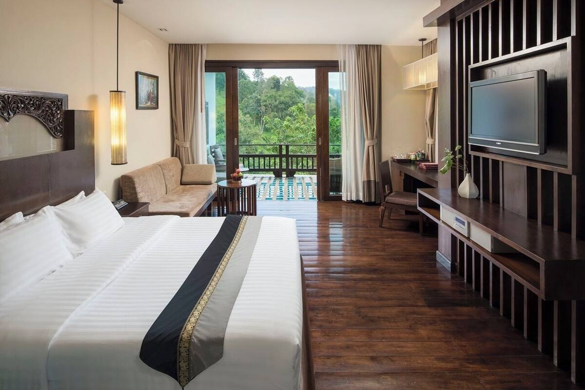 Quarto de hotel com uma cama grande, um sofá, uma escrivaninha com televisão e varanda com vista para o verde.