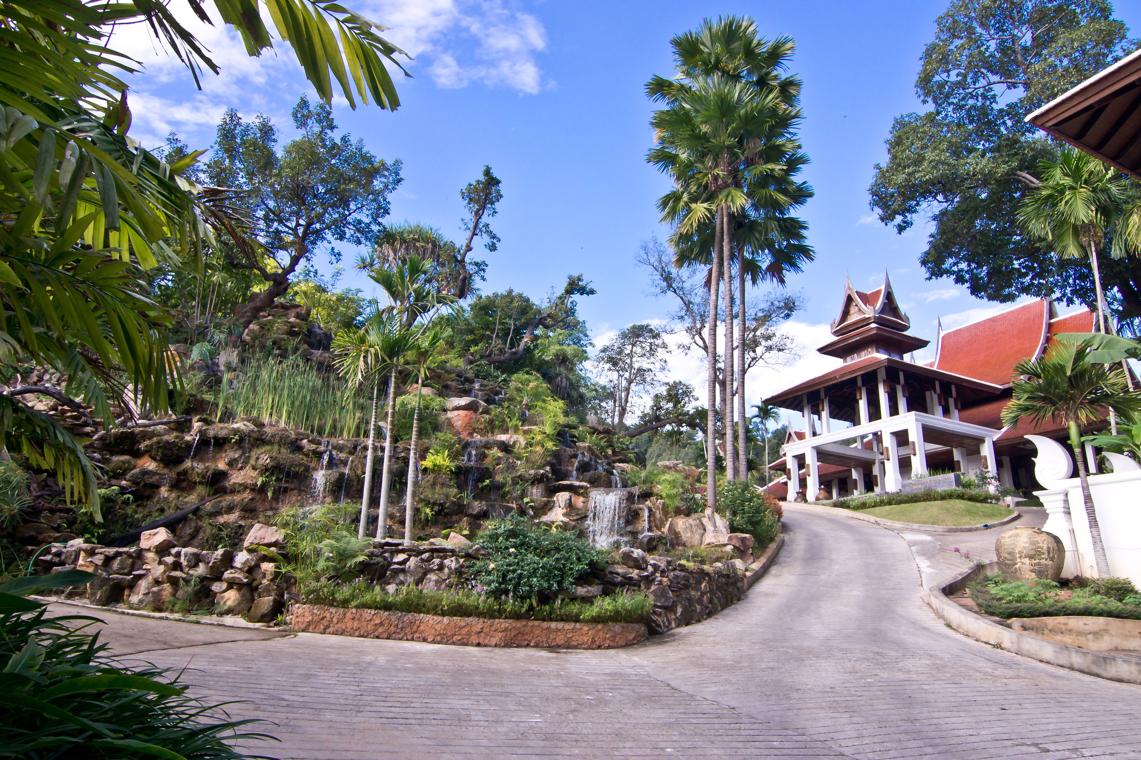 A paved road leading to a building with a traditional architectural style, surrounded by lush greenery and palm trees.