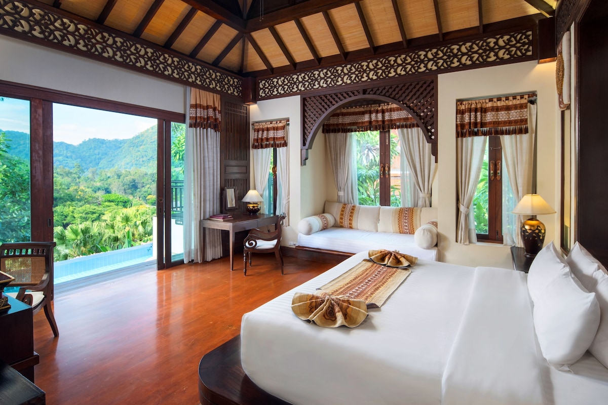 A hotel room with a large bed, a desk, and a view of lush greenery.