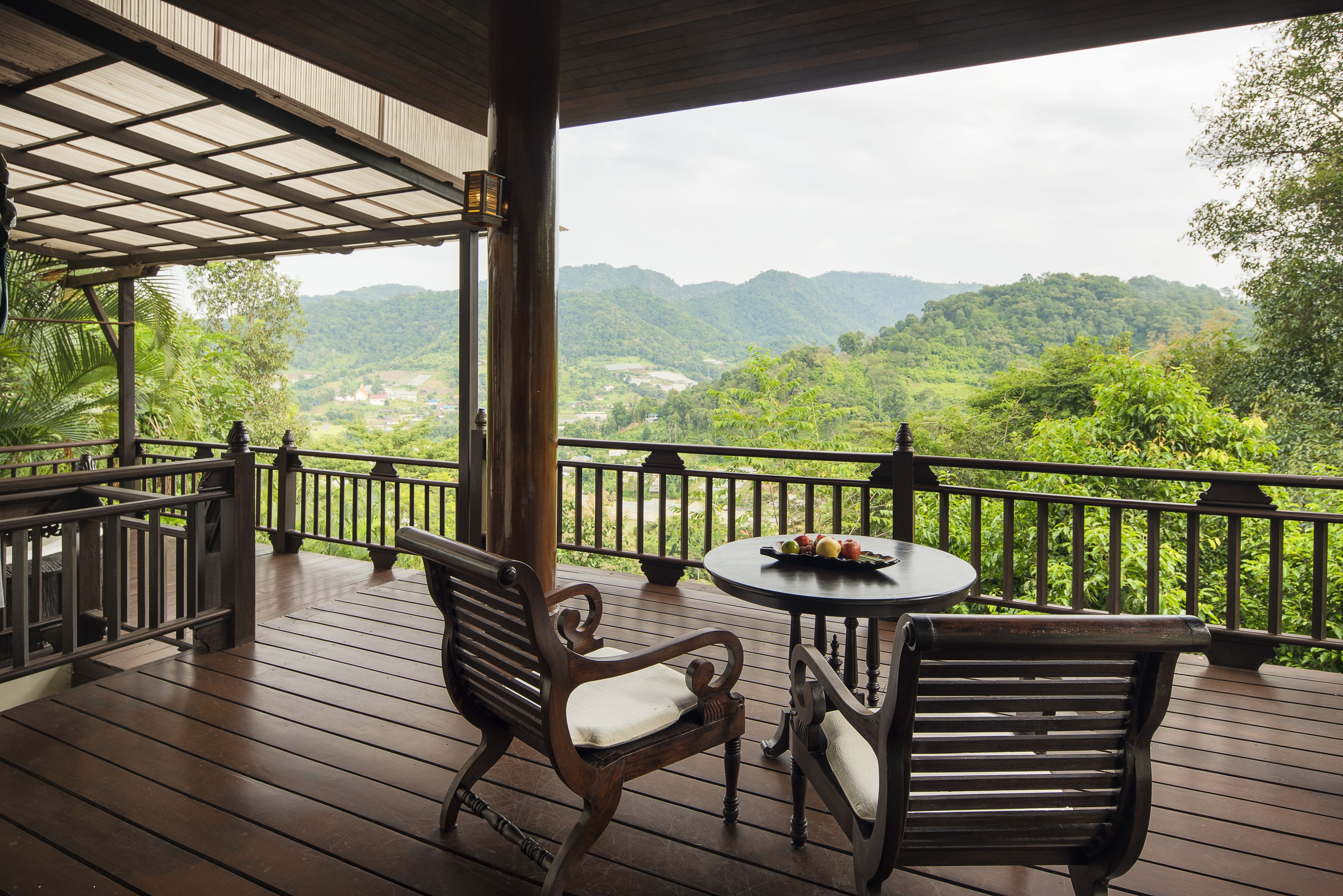 A wooden deck with two chairs and a table, overlooking a lush valley and mountains.