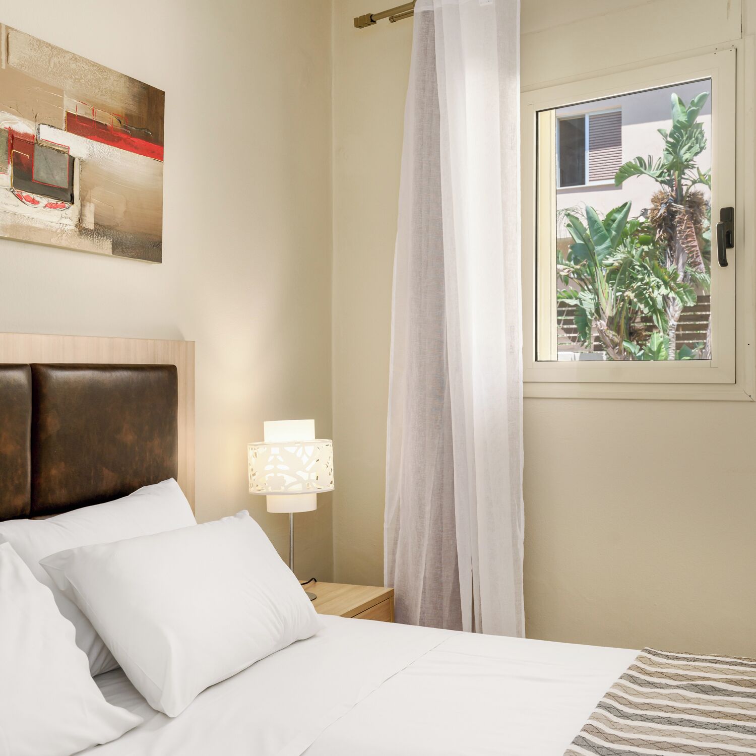 A hotel room with a bed, a nightstand, a lamp, a window with a view of greenery, and a painting on the wall.