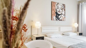 A hotel room with a large bed, two bedside lamps, a nightstand, a white chair, and a wall-mounted painting. - Axos Hotel (Rethymno)