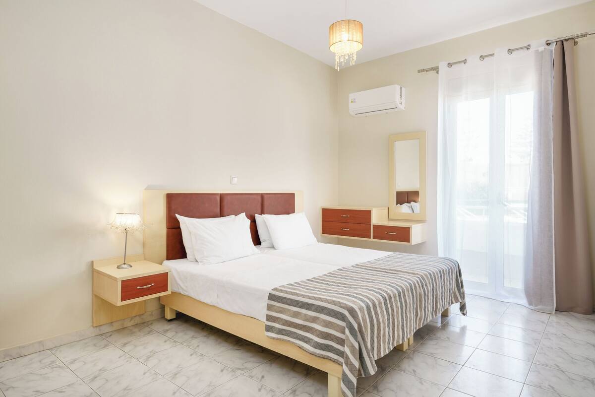 Ein Schlafzimmer mit Bett, Nachttisch, Kommode und einem großen Fenster mit Vorhängen.