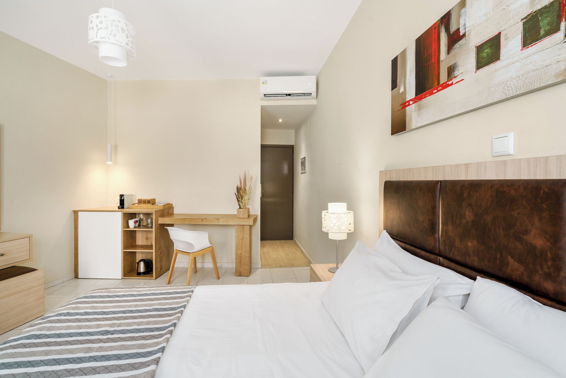 A modern hotel room with a large bed, a desk with a chair, a small kitchenette, and a large window.