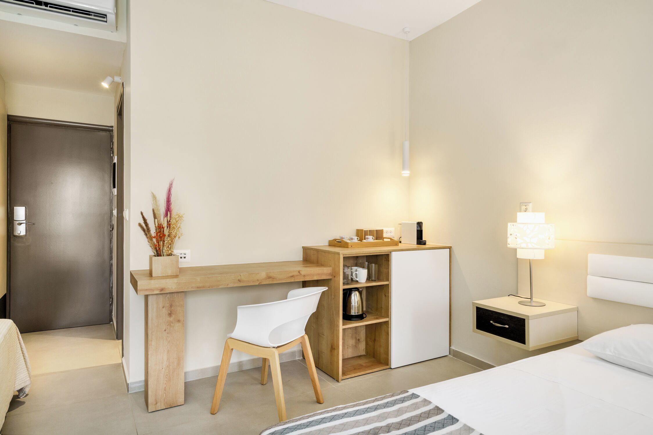 A modern hotel room with a wooden desk, white chair, bedside table with lamp, and a bed.