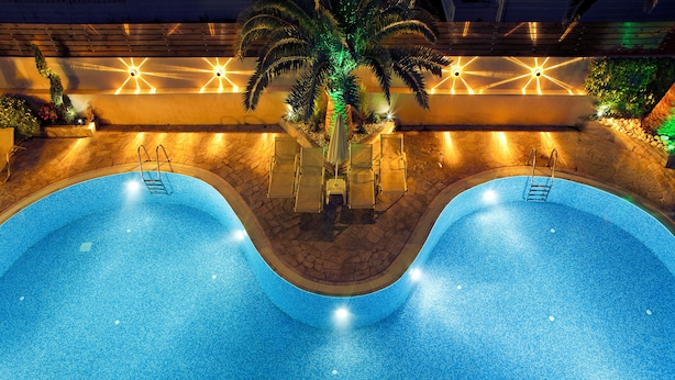 A swimming pool with two separate sections, surrounded by lounge chairs and a palm tree. - Axos Hotel (Rethymno)