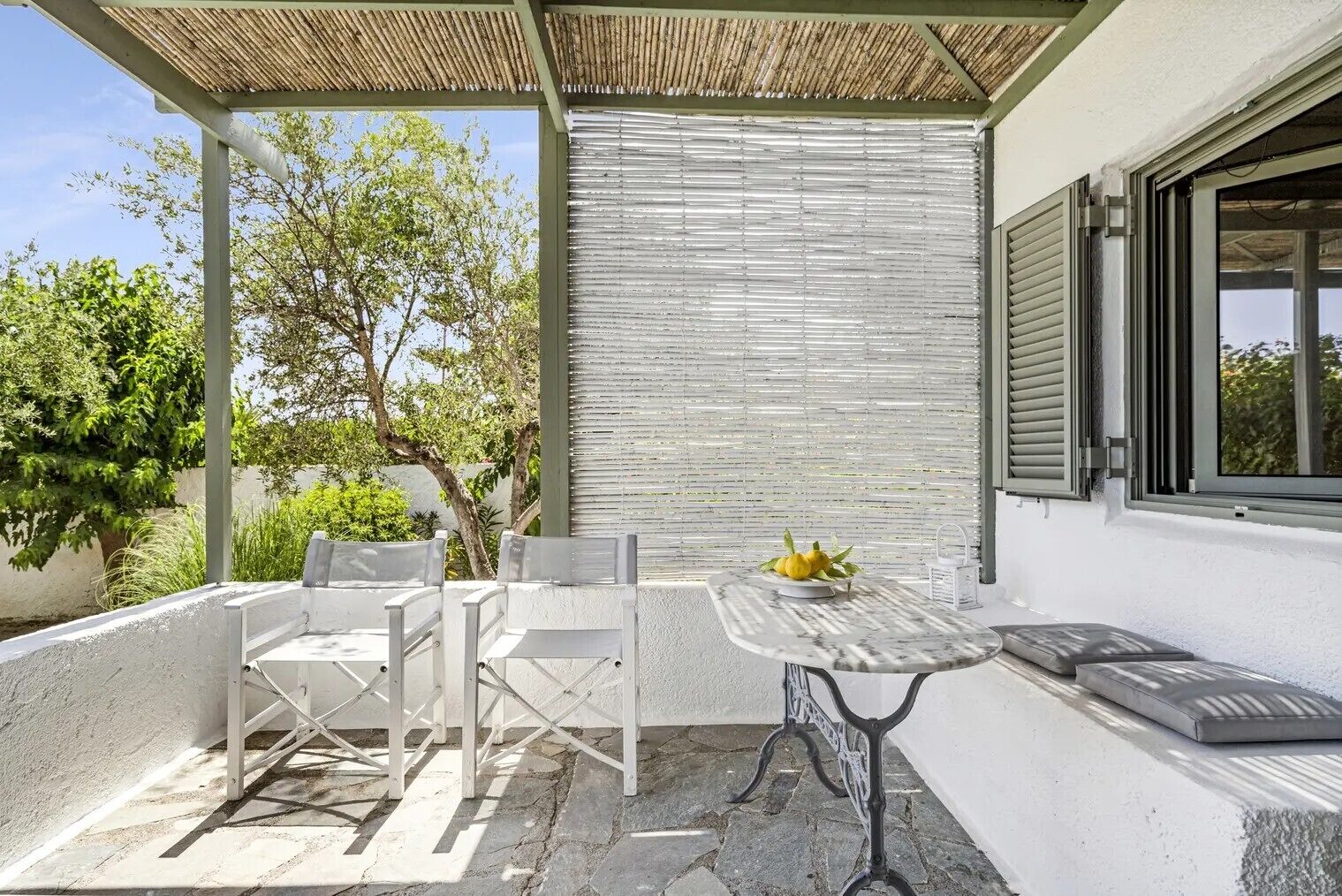 Un patio al aire libre con mesa y sillas, una pérgola y vistas a árboles y un edificio.