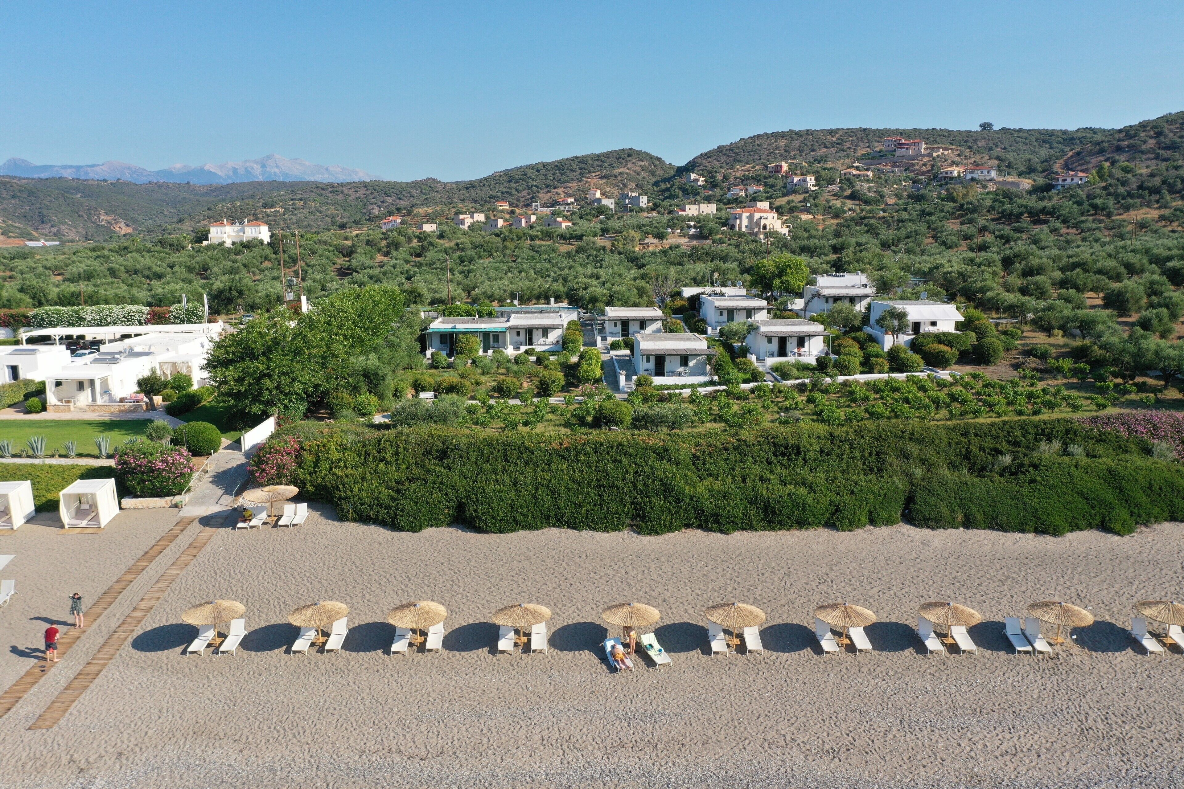 A resort with multiple buildings, a beach with sun loungers and umbrellas, and a mountainous backdrop.