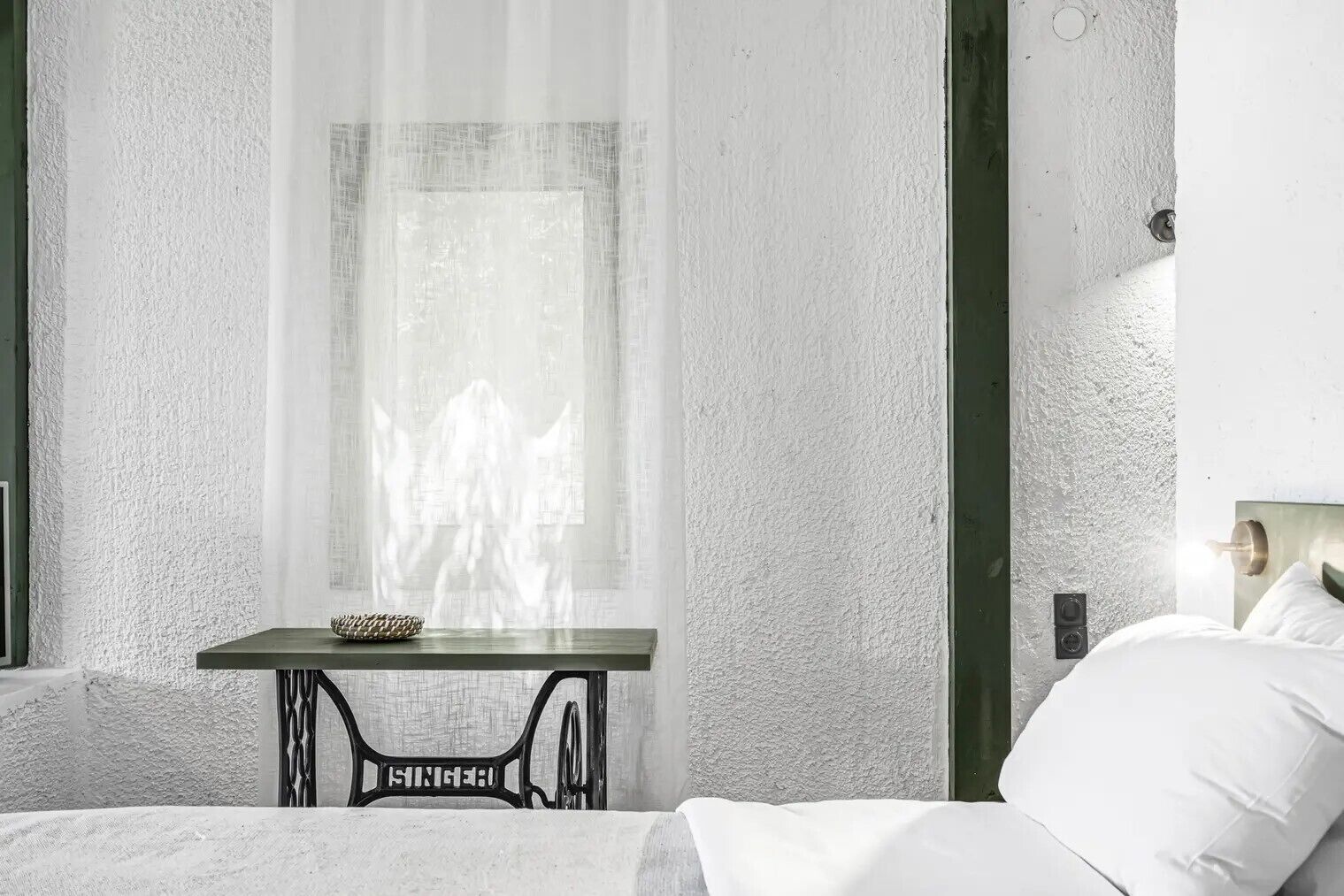 A bedroom with a bed, a small table with a lamp, a window with sheer curtains, and a textured wall.