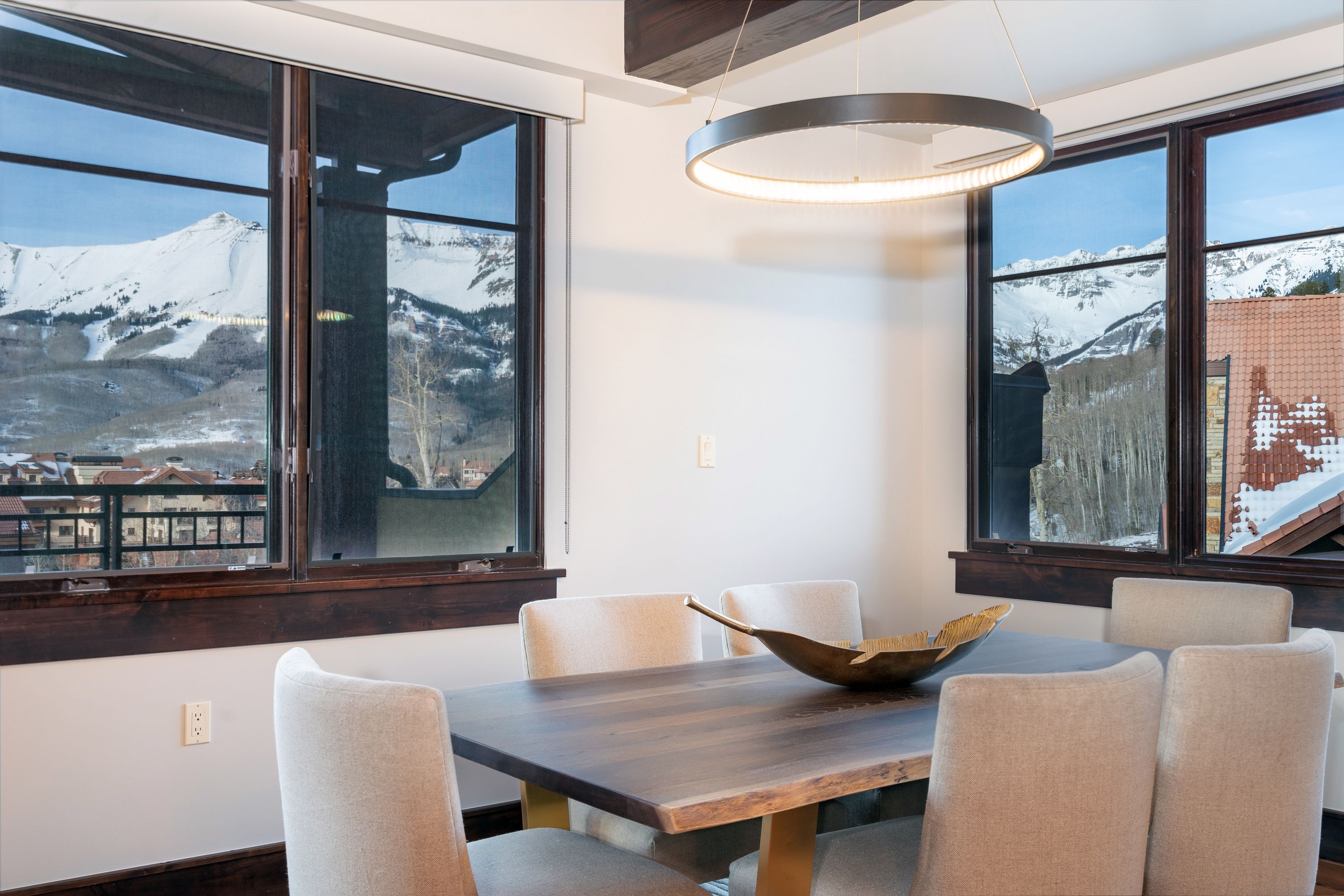 A dining area with a wooden table and six chairs, large windows with a view of snow-capped mountains, and a modern pendant light.