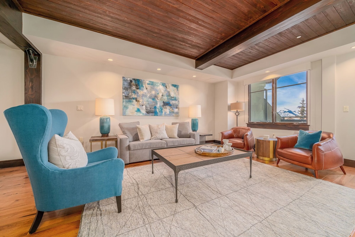 A spacious living room with a high ceiling, a large abstract painting, a sofa, two armchairs, a coffee table, and a window with a mountain view.