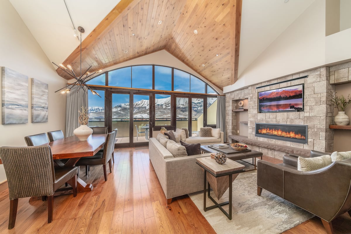 A spacious living area with a fireplace, large windows, and a high ceiling.