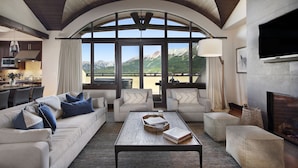 A spacious living room with a high ceiling, large windows offering mountain views, a fireplace, and a modern kitchen in the background. - Relais & Châteaux Lumière by Dunton (Telluride)
